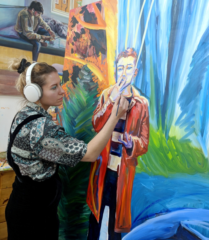 Julia is in her studio, wearing headphones and painting a large, colourful canvas of a young person who is smoking.